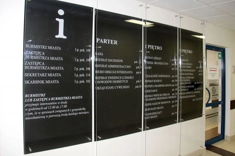 Tablice informacyjne z nazwami urzędów i numerami pokoi umieszczone na ścianie korytarza.